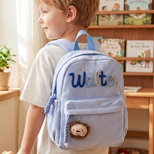 May include: A light blue corduroy backpack featuring the name Walter embroidered in dark blue script on the front pocket. The bag includes a small, plush lion face patch attached to the lower section and a braided cord zipper pull detail.