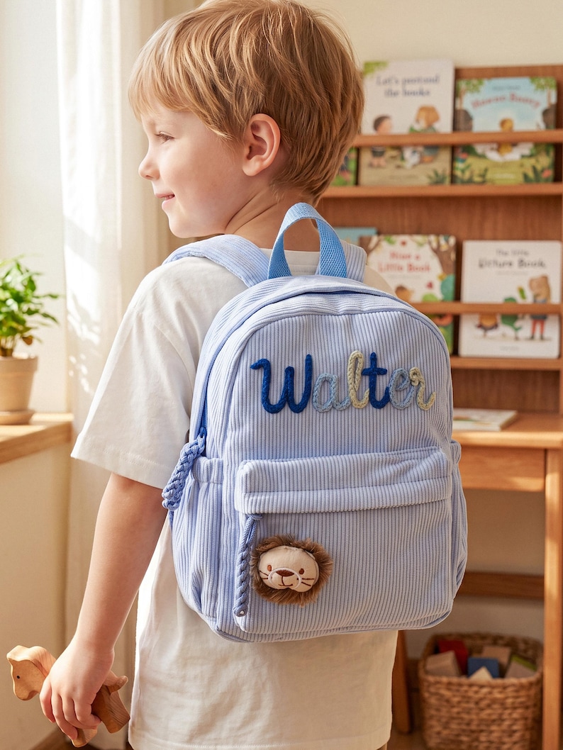 May include: A light blue corduroy backpack featuring the name Walter embroidered in dark blue script on the front pocket. The bag includes a small, plush lion face patch attached to the lower section and a braided cord zipper pull detail.