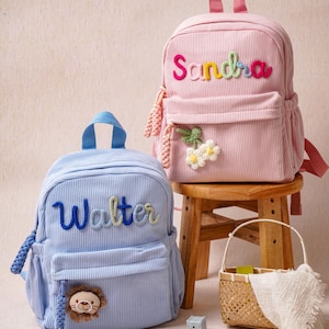 May include: Two corduroy backpacks, one light blue and one light pink, each featuring a top handle and a front pocket. The blue backpack displays the name Walter in blue yarn and a lion patch. The pink backpack shows the name Sandra in multicolored yarn and a white flower patch.