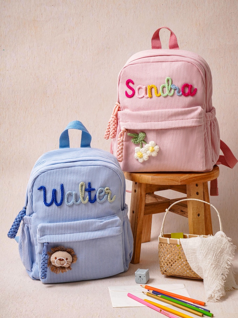 May include: Two corduroy backpacks, one light blue and one light pink, each featuring a top handle and a front pocket. The blue backpack displays the name Walter in blue yarn and a lion patch. The pink backpack shows the name Sandra in multicolored yarn and a white flower patch.