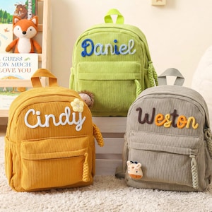 May include: Three corduroy backpacks in lime green, mustard yellow, and gray. The lime green bag features the name Daniel in blue. The mustard yellow bag displays the name Cindy in white with a flower. The gray bag shows the name Weston in brown.