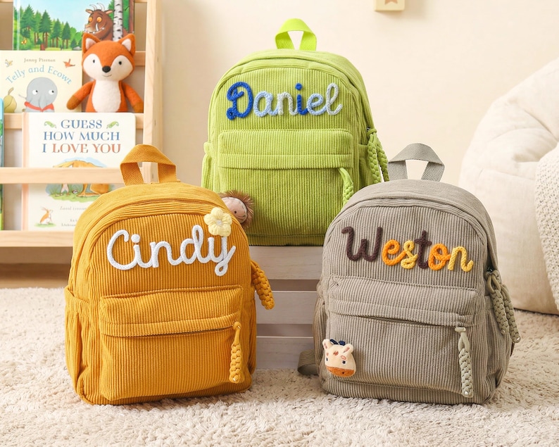 May include: Three corduroy backpacks in lime green, mustard yellow, and gray. The lime green bag features the name Daniel in blue. The mustard yellow bag displays the name Cindy in white with a flower. The gray bag shows the name Weston in brown.