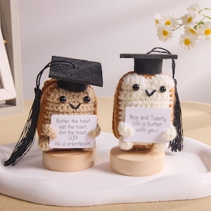 Puede incluir: Dos figuras de tostadas de ganchillo con birretes. Una sostiene un letrero que dice "Butter the toast, eat the toast, shit the toast, GOD! life is erlentless!!!" La otra dice "Nice and Toasty life is butter with you!"