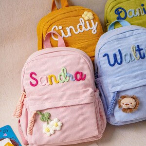 May include: A collection of corduroy backpacks in pastel and bright colors, each featuring a personalized name embroidered in yarn. The pink bag displays the name Sandra with a flower applique, while the blue bag features the name Walter and a lion face.