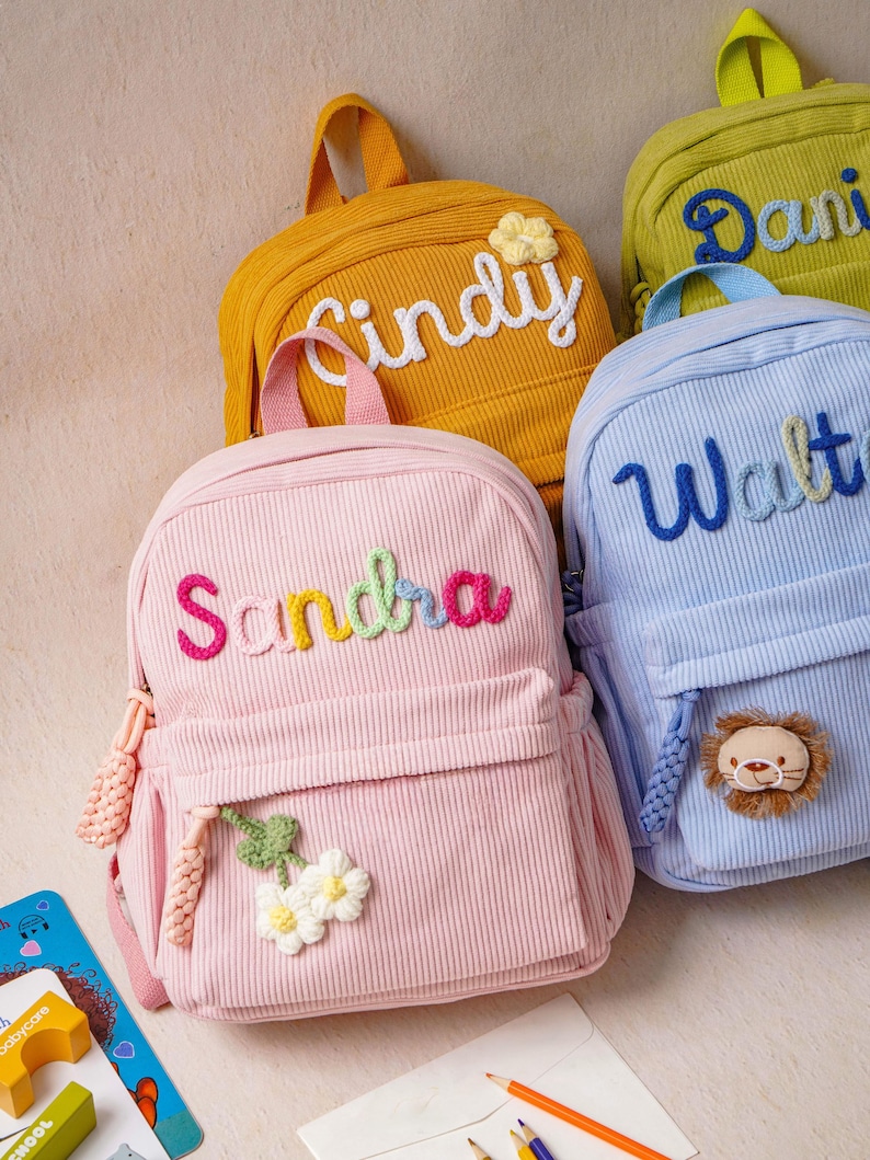 May include: A collection of corduroy backpacks in pastel and bright colors, each featuring a personalized name embroidered in yarn. The pink bag displays the name Sandra with a flower applique, while the blue bag features the name Walter and a lion face.