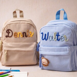 May include: Two corduroy backpacks, one beige and one light blue, each featuring a top handle, a front zippered pocket, and side pockets. The beige backpack has the name Dennis embroidered in brown and yellow, while the blue one says Walter in blue.