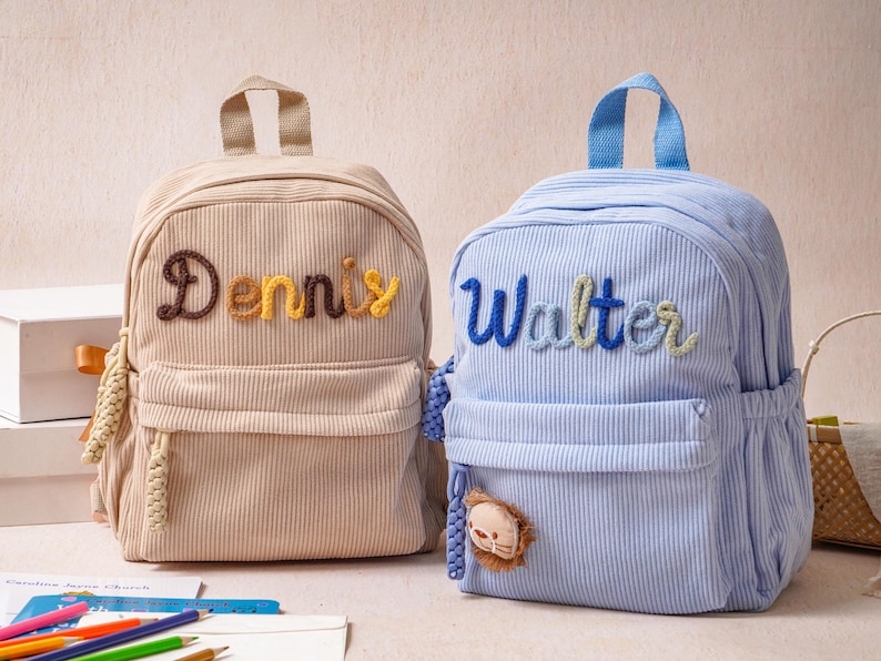 May include: Two corduroy backpacks, one beige and one light blue, each featuring a top handle, a front zippered pocket, and side pockets. The beige backpack has the name Dennis embroidered in brown and yellow, while the blue one says Walter in blue.