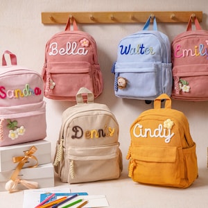 May include: A collection of six corduroy backpacks in various colors including pink, light blue, beige, and yellow. Each backpack features a personalized name embroidered on the front, along with small decorative floral or animal-shaped accents.