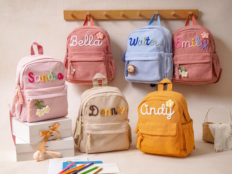 May include: A collection of six corduroy backpacks in various colors including pink, light blue, beige, and yellow. Each backpack features a personalized name embroidered on the front, along with small decorative floral or animal-shaped accents.