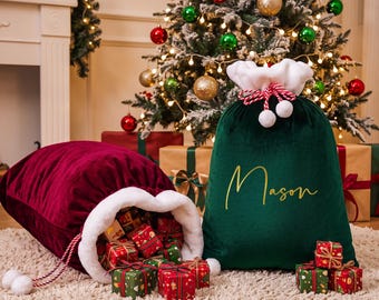 May include: Two velvet Christmas gift sacks with white faux fur trim and red and white braided drawstring cords. One sack is deep red and lying on its side, while the other is dark green with the name Mason embroidered in gold script.