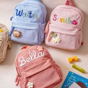 May include: Three corduroy backpacks in light blue, light pink, and dusty rose. Each bag features a top handle, a front zippered pocket, and embroidered names: Walter, Sandra, and Bella. Decorative crochet appliques include a lion and flowers.