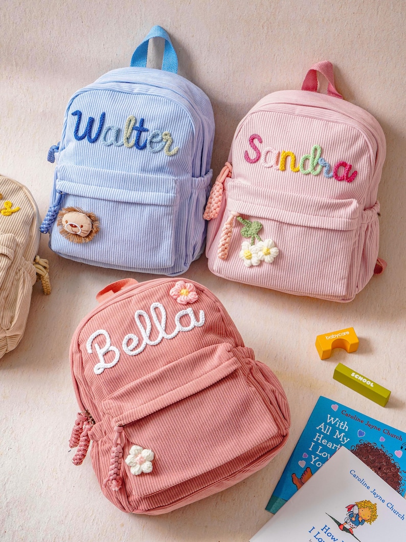 May include: Three corduroy backpacks in light blue, light pink, and dusty rose. Each bag features a top handle, a front zippered pocket, and embroidered names: Walter, Sandra, and Bella. Decorative crochet appliques include a lion and flowers.