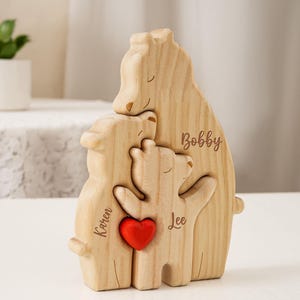 Könnte beinhalten: Holzfigur einer Bärenfamilie, die sich umarmt. Die Bären sind aus hellem Holz gefertigt und tragen die Namen "Bobby", "Karen" und "Lee". Ein rotes Herz befindet sich in der Mitte. Im Hintergrund eine kleine Pflanze in einem weißen Topf.