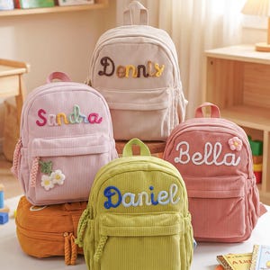 May include: Four corduroy backpacks in beige, light pink, lime green, and dusty rose. Each bag features a front pocket, a top handle, and a personalized name embroidered in colorful yarn. The pink bag has white flower accents, and the rose bag has a flower.