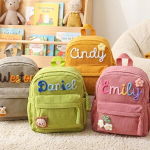 May include: Four corduroy backpacks in gray, green, yellow, and pink. Each bag features a top handle, a front zippered pocket, and a personalized name embroidered in yarn. The bags are decorated with small plush animal or flower appliques.