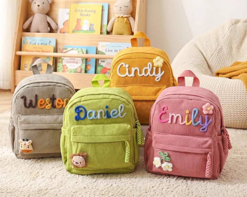 May include: Four corduroy backpacks in gray, green, yellow, and pink. Each bag features a top handle, a front zippered pocket, and a personalized name embroidered in yarn. The bags are decorated with small plush animal or flower appliques.