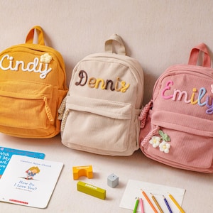 May include: Three corduroy backpacks in mustard yellow, beige, and dusty pink. Each bag features a front pocket, a top handle, and embroidered names: Cindy, Dennis, and Emily. The pink bag includes small flower appliqu&eacute;s, while the yellow bag has one.