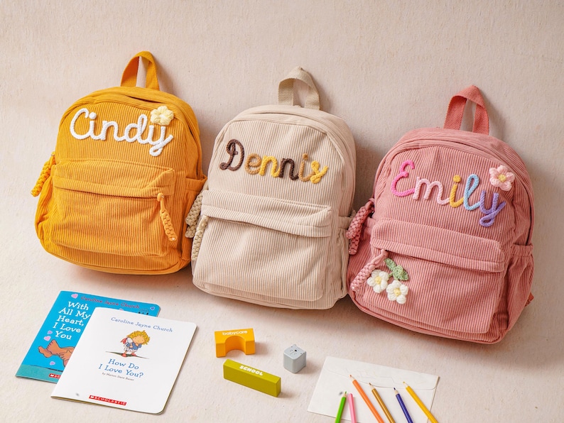 May include: Three corduroy backpacks in mustard yellow, beige, and dusty pink. Each bag features a front pocket, a top handle, and embroidered names: Cindy, Dennis, and Emily. The pink bag includes small flower appliqu&eacute;s, while the yellow bag has one.