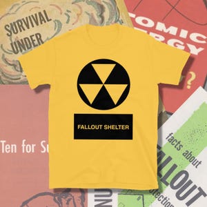 May include: Yellow t-shirt featuring a black radiation symbol inside a black circle, above a black rectangle with the words "FALLOUT SHELTER" in white. The shirt is a short-sleeved crew neck.