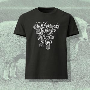 May include: Black crew neck t-shirt with the text "Do Androids Dream of Electric Sheep" in white, stylized script. The shirt is set against a light green background with a sheep illustration.