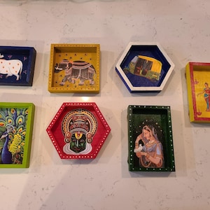 Puede incluir: Colección de siete bandejas de madera pintadas a mano y coloridas. Cada bandeja presenta un diseño único, que incluye una vaca, un elefante, un pavo real y figuras. Las bandejas tienen varias formas, incluyendo rectangulares y hexagonales.