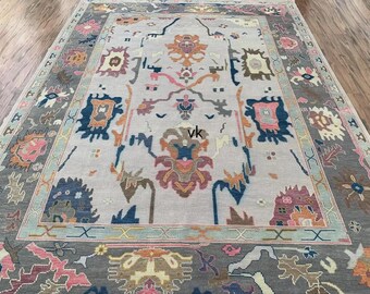 May include: A large, rectangular area rug with an intricate, colorful design. The rug features a light gray central field with a border in shades of gray, blue, pink, and orange. The design includes floral and geometric patterns.