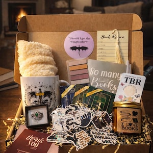 Puede incluir: Una caja de regalo llena de artículos temáticos de libros. Incluye una taza, pegatinas, un anillo, una vela y una tarjeta de reseña de libro. La caja contiene una variedad de artículos, incluyendo una taza con un diseño de calavera y un anillo con una piedra verde.