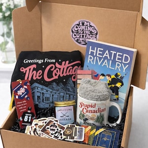 Peut inclure: Une boîte cadeau marron remplie d'articles sur le thème du chalet. Comprend un t-shirt noir avec le texte "Greetings From The Cottage", un livre intitulé "Heated Rivalry", une tasse, une bougie, des autocollants et un marque-page. La boîte porte un autocollant "I'm Coming to the Cottage".