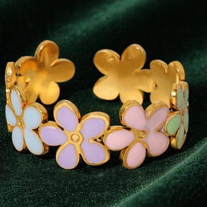 May include: A gold-toned bracelet featuring a floral design. The bracelet is adorned with alternating pastel-colored flower petals in shades of white, lavender, pink, and green. The bracelet is set against a dark green velvet background.