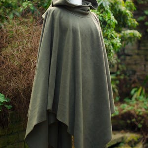 May include: A hooded green fleece poncho with a high neck and a long, flowing design. The poncho is displayed on a wooden stand against a backdrop of green foliage.