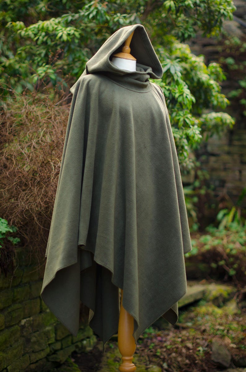 May include: A hooded green fleece poncho with a high neck and a long, flowing design. The poncho is displayed on a wooden stand against a backdrop of green foliage.