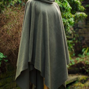 May include: A hooded green fleece poncho with a long, flowing design. The poncho is displayed on a wooden mannequin against a backdrop of green foliage.
