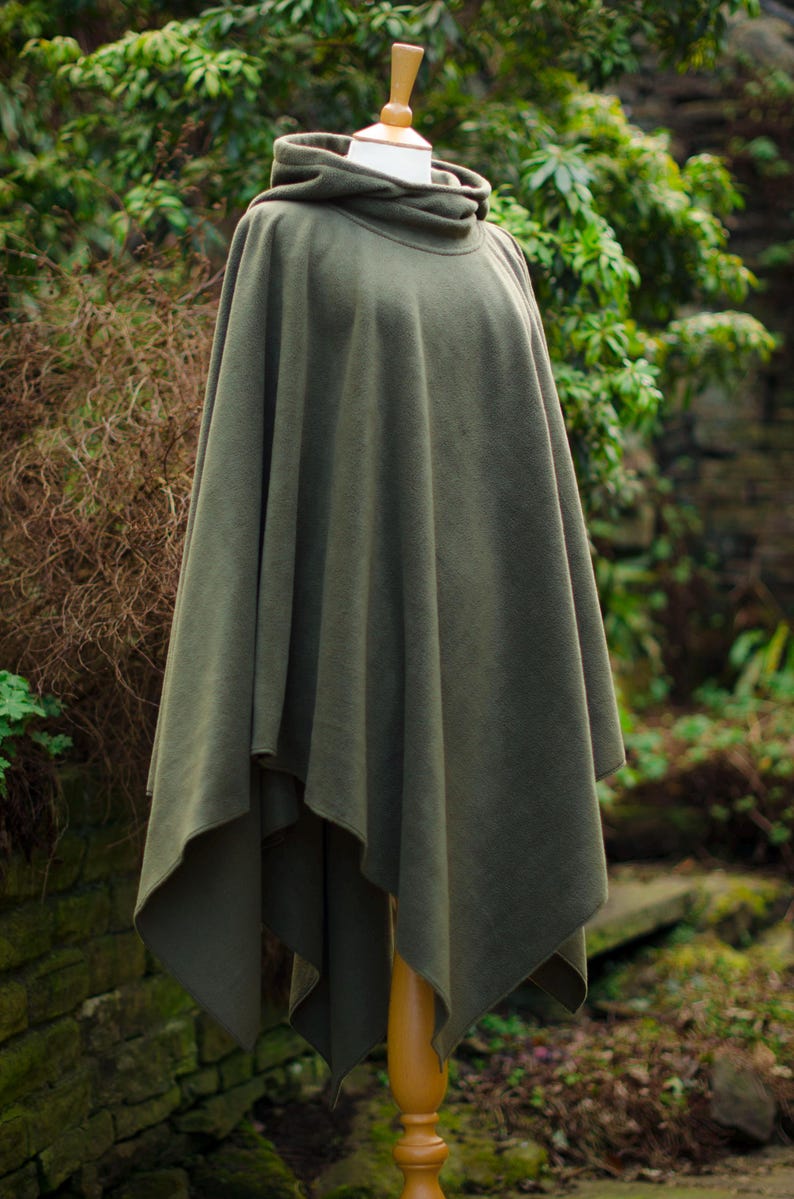May include: A hooded green fleece poncho with a long, flowing design. The poncho is displayed on a wooden mannequin against a backdrop of green foliage.