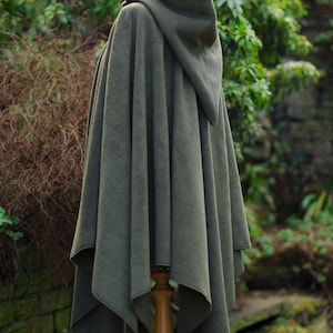 May include: A hooded green fleece cape with a pointed hood. The cape is draped over a wooden mannequin and is displayed against a backdrop of green foliage.