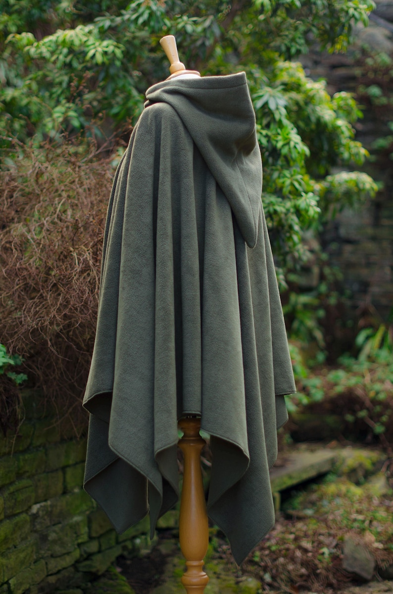May include: A hooded green fleece cape with a pointed hood. The cape is draped over a wooden mannequin and is displayed against a backdrop of green foliage.