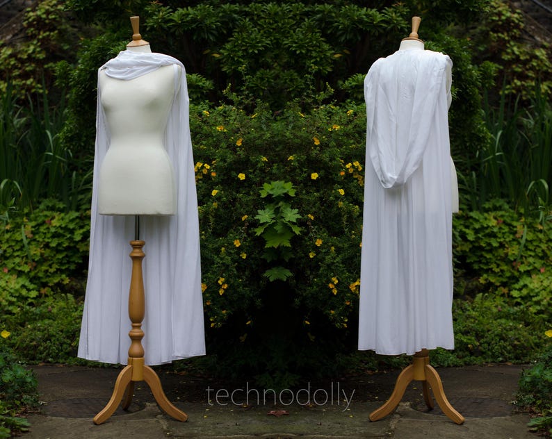 Pode incluir: Duas capas brancas expostas em manequins. Uma capa tem capuz, a outra tem cachecol. Os manequins est&atilde;o em suportes de madeira. O fundo &eacute; um exuberante jardim verde. O texto "technodolly" &eacute; vis&iacute;vel.