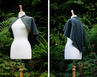 May include: A dark green hooded cape with a simple design, displayed on a white mannequin against a backdrop of lush green foliage.