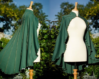 May include: A green hooded cape with gold trim and a scalloped edge. The cape is displayed on a mannequin against a backdrop of green foliage.