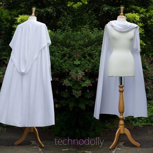 Pode incluir: Duas capas brancas em manequins de madeira. Uma capa tem capuz, a outra tem gola drapeada. Os manequins est&atilde;o em suportes de madeira. O fundo &eacute; um jardim verde com o texto 'technodolly'.