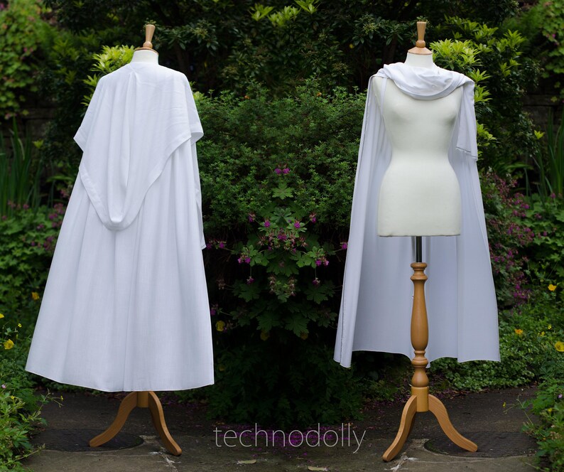 Pode incluir: Duas capas brancas em manequins de madeira. Uma capa tem capuz, a outra tem gola drapeada. Os manequins est&atilde;o em suportes de madeira. O fundo &eacute; um jardim verde com o texto 'technodolly'.