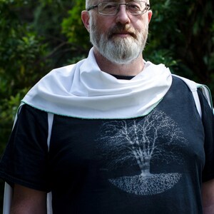 Pode incluir: Uma pessoa vestindo uma camiseta preta com um gr&aacute;fico de &aacute;rvore branca. Ele tamb&eacute;m est&aacute; usando um cachecol branco e &oacute;culos. O fundo &eacute; uma folhagem verde desfocada.