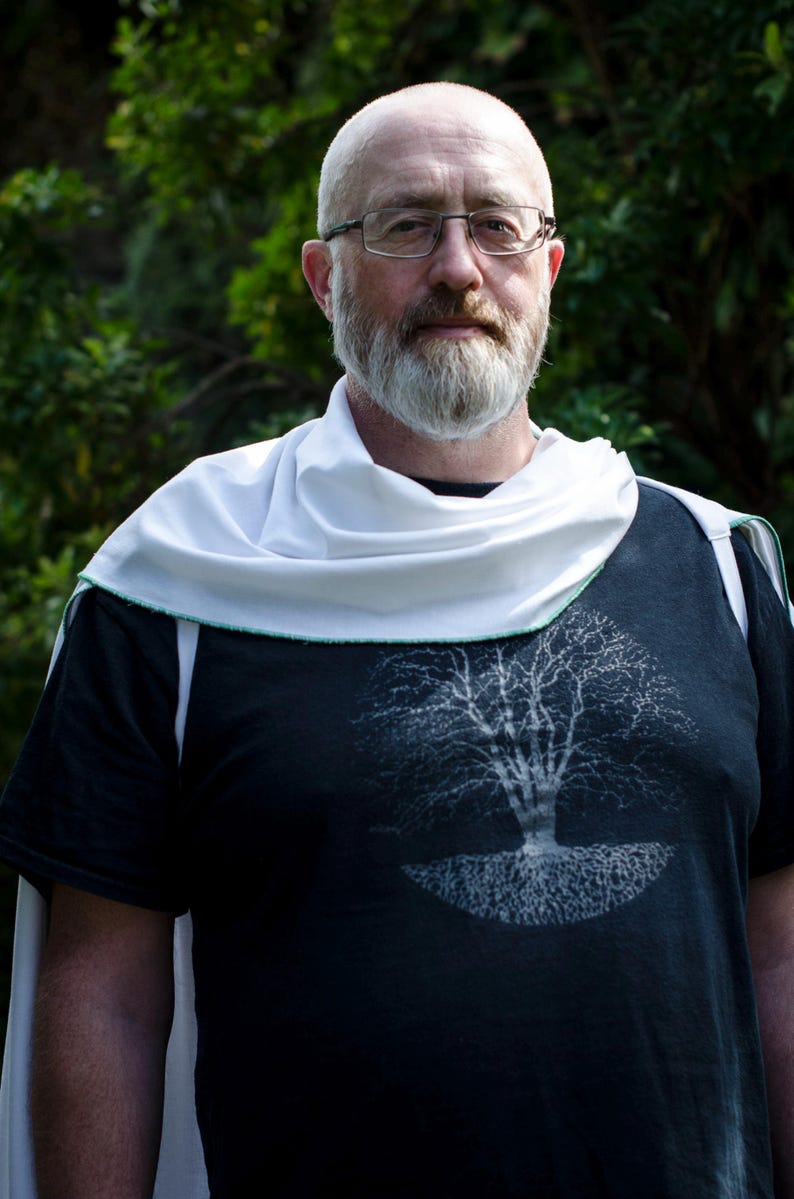 Pode incluir: Uma pessoa vestindo uma camiseta preta com um gr&aacute;fico de &aacute;rvore branca. Ele tamb&eacute;m est&aacute; usando um cachecol branco e &oacute;culos. O fundo &eacute; uma folhagem verde desfocada.