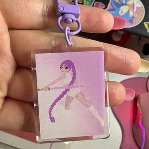 Puede incluir: Un llavero acrílico transparente con un cierre morado. El dije rectangular presenta un fondo degradado brillante y una ilustración de una guerrera con una espada. La guerrera tiene pelo largo y trenzado morado y viste ropa blanca.