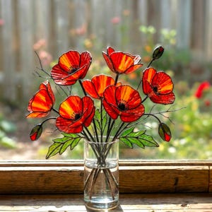 Puede incluir: Un jarrón de cristal con un ramo de amapolas rojas y naranjas con hojas verdes. Las flores están frente a una ventana con un alféizar de madera. La sombra de las flores se proyecta sobre la superficie de madera.
