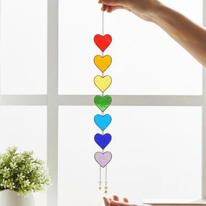 May include: A rainbow-colored heart-shaped suncatcher. The suncatcher features seven hearts in a vertical line, each a different color of the rainbow. The hearts are suspended by a string with decorative beads.