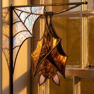 Pode incluir: Um apanhador de sol em vitral com um morcego pendurado numa haste de metal, com vidro branco iridescente a formar um padrão de teia de aranha no canto e vidro cor de âmbar para o corpo e asas, unidos por linhas de soldadura de metal escuro.