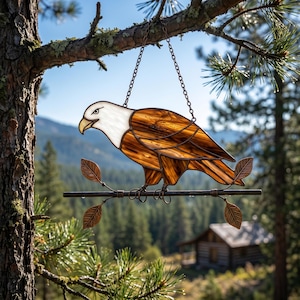 Könnte beinhalten: Glasfenster-Adler-Suncatcher mit weißem Kopf und braunem Körper, der auf einem Metallzweig mit bronzefarbenen Blättern sitzt. Der Adler hängt an einer Kette, perfekt für die Außendekoration. Im Hintergrund eine Hütte und eine Berglandschaft.