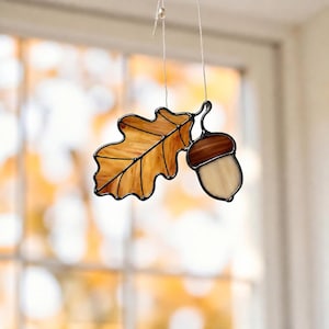 May include: A stained glass suncatcher featuring an amber-colored oak leaf attached to a brown and cream-colored acorn. The piece is constructed with dark metal solder lines and hangs from a thin white string against a blurred window background.