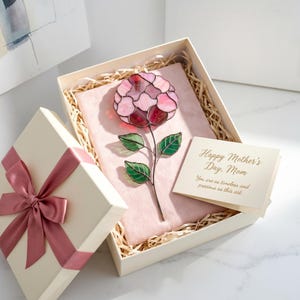 May include: A decorative stained glass flower in a cream gift box with a pink ribbon. The flower is pink and red with green leaves. A card inside the box reads "Happy Mother's Day, Mom". The box is filled with straw.
