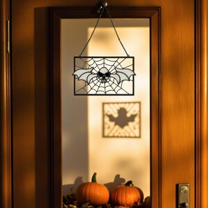May include: A Halloween decoration with a black frame and a spiderweb design. A skull with bat wings is in the centre of the web. The decoration hangs from a chain on a wooden door. Two orange pumpkins are visible below, adding to the autumnal theme.